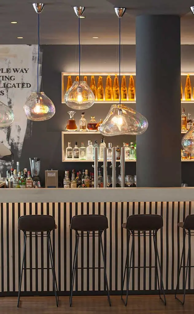 Bar mit Lamellenfront, Barhockern und Glasleuchten vor einem Flaschenregal